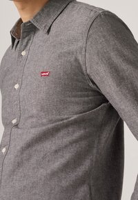 Graues Hemd mit langen Ärmeln, feiner Textur, mit einem roten "Levi's"-Logo auf der Brust und standardmäßigen weißen Knöpfen.