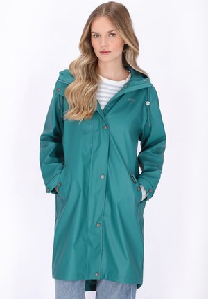 Femme aux longs cheveux blonds ondulés portant un imperméable teal boutonné, mains dans les poches, sur une chemise rayée et un jean bleu.