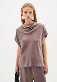 Top corto e morbido di colore malva con scollatura drappeggiata. La texture è morbida e leggermente stropicciata, in armonia con pantaloni dalla vestibilità rilassata.