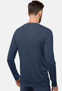 Homme aux cheveux courts et foncés portant une chemise unie bleu marine à manches longues, vu de dos contre un fond gris.