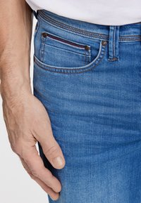 Męskie niebieskie dżinsy o gładkiej fakturze, z przednią kieszenią z czerwono-białym akcentem, miedzianymi nitami i klasycznym krojem.