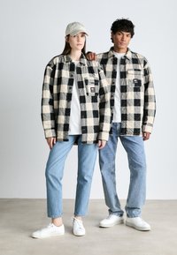 Du šie asmenys dėvi dideles languotas marškinėlius juoda ir grietinėlės spalvos, derindami su baltais marškiniais ir džinsais. Jie stovi ant neutralios grindų.