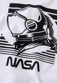 Baltas marškinėlis su juodu astraunauto šalmą vaizduojančiu piešiniu ir žodžiu „NASA“, fone horizontalios linijos, sukuriantys dinamišką efektą.
