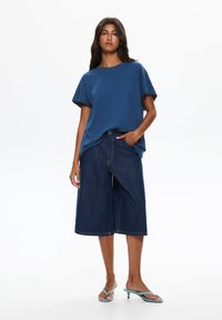 T-shirt di cotone blu con maniche corte, abbinato a culotte di denim scuro. La modella indossa sandali con tacco verde. Design semplice e vestibilità rilassata.