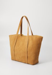 Sac fourre-tout en daim beige avec deux longues poignées, forme carrée, détails de couture visibles et un design simple. Texture lisse avec des accents minimalistes.