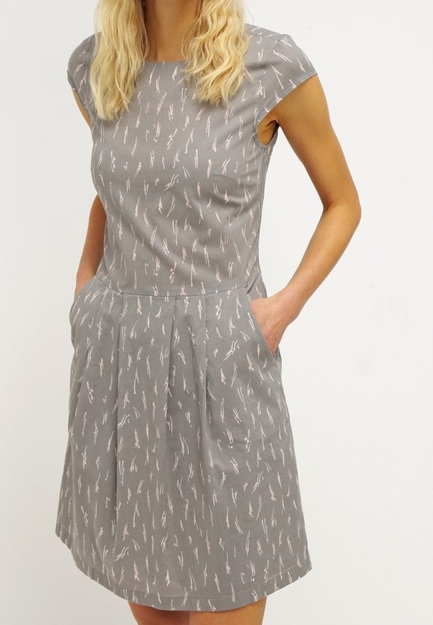 Femme aux cheveux blonds portant une robe grise à manches courtes avec des motifs abstraits blancs et les mains dans les poches latérales.