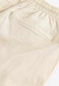 Pantalons beiges avec une taille élastique, dotés d'une poche arrière et d'une texture lisse. Le tissu semble léger et légèrement réfléchissant.