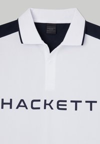 Polo bianco con colletto e dettagli sulle spalle in blu navy, caratterizzato da un logo "HACKETT" prominente in lettere scure. Tessuto testurizzato.