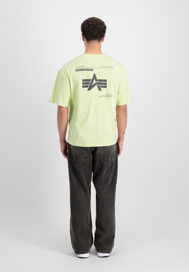 Camiseta verde lima con un gran diseño gráfico negro en la parte posterior, de manga corta, corte holgado, combinada con unos pantalones vaqueros sueltos de color gris oscuro y zapatillas blancas.