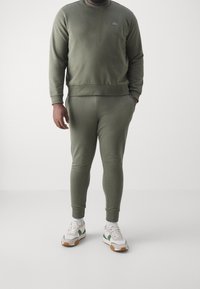 Bluza i spodnie dresowe w kolorze oliwkowej zieleni wykonane z miękkiego materiału, z okrągłym dekoltem i ściągaczami na rękawach, zestawione z białymi i zielonymi butami sportowymi.