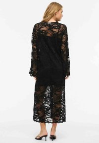 Femme portant une longue robe noire en dentelle florale à manches longues et des sandales noires à talons, vue de dos sur fond blanc.