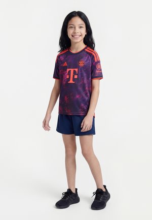 Lachend meisje met lang zwart haar, gekleed in een paars-oranje sportshirt, marineblauwe korte broek en zwarte sneakers, staand tegen een witte achtergrond.