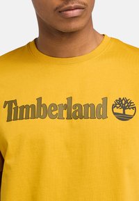 Keltainen puuvillapaita, jossa on tummanharmaa "Timberland" -logo ja puun symboli. Paita sisältää klassisen pyöreän pääntien ja lyhyet hihat.