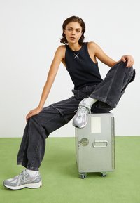 Giovane che indossa una canotta nera e pantaloni larghi grigi, seduto su una valigia metallica argentata con pavimento verde e sfondo bianco.