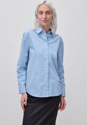 Vrouw met lichtblond haar die een blauw-wit gestreept overhemd met knopen en een donkere fijn gestreepte rok draagt tegen een effen achtergrond.