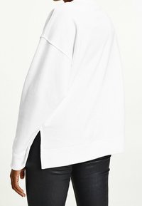 Persona che indossa una felpa bianca larga con fessure laterali e pantaloni neri, mostrata di spalle dalla vita fino a metà coscia.