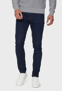 Hombre que lleva unos pantalones vaqueros slim-fit de color azul oscuro, un suéter texturizado gris claro y zapatillas blancas, de pie contra un fondo liso claro.