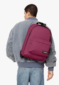 Mochila burdeos con un bolsillo frontal, cierre de cremallera y etiqueta. Fabricada con un tejido duradero, cuenta con correas para los hombros y un diseño elegante y redondeado.