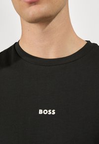 Černé bavlněné tričko s kulatým výstřihem a bílým logem značky "BOSS" umístěným na hrudi. Hladká textura, minimalistický design.