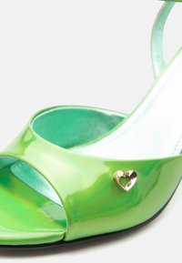 Πράσινο παπούτσι από δερμάτινο λάκα με ανοιχτό δάχτυλο και ψηλό τακούνι, με μικρό μεταλλικό στολίδι σε σχήμα καρδιάς στο πλάι.