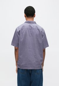 Rövid ujjú ing világoslila, texturált anyagból. Gallérral és hátrészével, laza szabású kék farmerral párosítva.