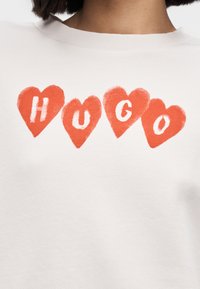 Bela majica s kapuco z izrazitimi rdečimi srci in besedo "HUGO" v teksturirani, igrivi pisavi. Rebrast ovratnik dodaja detajl.