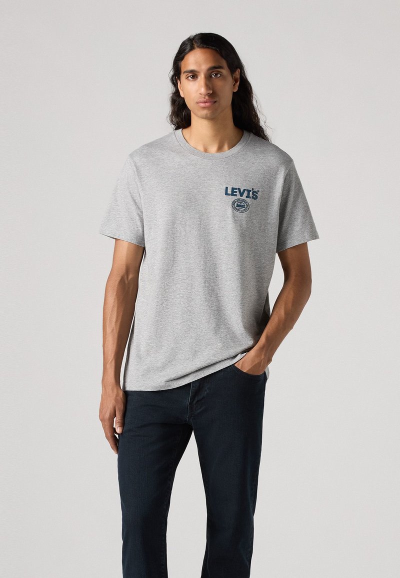 Camiseta de algodón gris con mangas cortas y escote redondo. Presenta un logo azul de Levi's en el pecho. Combinada con jeans oscuros.