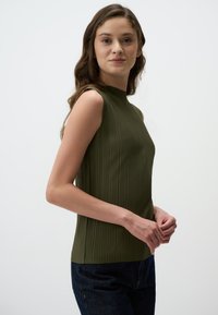 Top senza maniche di colore verde khaki con pieghe verticali, design a collo alto e silhouette aderente, abbinato a jeans scuri.