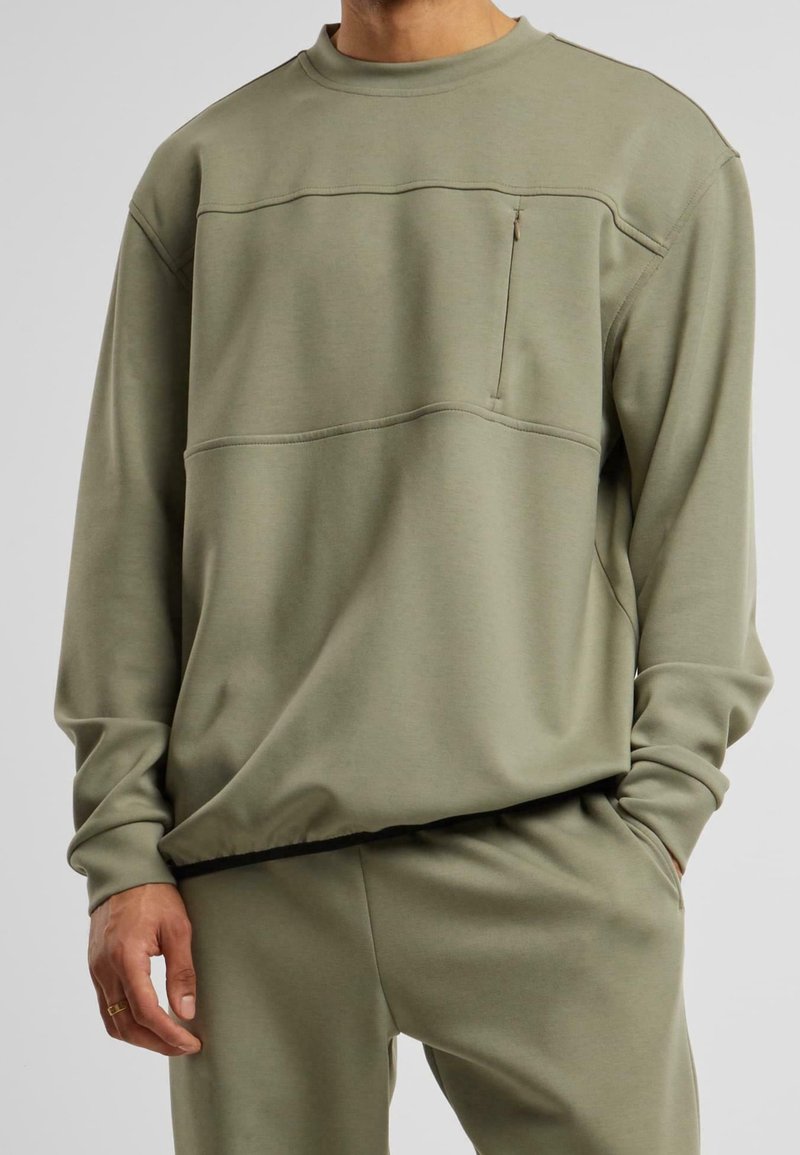 Sweat-shirt vert olive clair avec col rond, poche visible sur la poitrine et panneaux horizontaux cousus. Texture lisse et coupe décontractée.