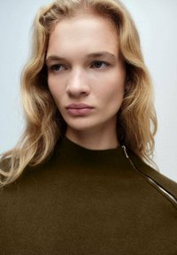 Maglia verde oliva alta con dettaglio zip sulla spalla, realizzata in tessuto a maglia, con una texture liscia e un design minimalista.