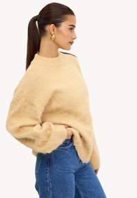 Sweater beige duveteux avec un col montant et des épaules tombantes, associé à un jean bleu taille haute. La texture semble douce et confortable.
