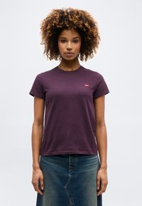 Γυναίκα με σγουρά μαλλιά που φοράει σκούρο μοβ κοντομάνικο μπλουζάκι και μπλε τζιν φούστα, στέκεται μπροστά σε απλό λευκό φόντο.