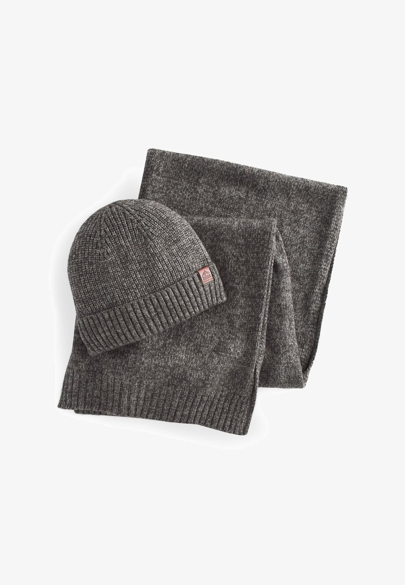 Szary zestaw czapki i szalika z dzianiny. Czapka ma prążkowany mankiet i małą łatkę z logo. Szalik wykonany jest z teksturowanej, miękkiej tkaniny.