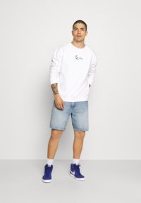 Witte sweatshirt met zwart logo, lichtblauwe denim shorts en blauwe high-top sneakers. Huid met zichtbare tatoeages op de arm. Neutrale achtergrond.