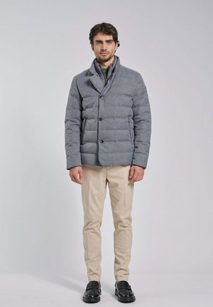 Man met een grijs gewatteerd jack, beige broek en zwarte schoenen, staand tegen een effen lichte achtergrond.