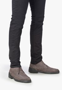 Botines de ante marrón con suelas de goma negras, que presentan una punta redondeada, cordones y una textura suave. Llevados con pantalones negros ajustados.