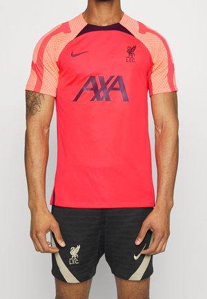 Mand iført rød og ferskenfarvet Liverpool FC fodboldtrøje med korte ærmer og sorte shorts med Liverpool-logo og Nike-swoosh.