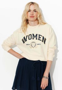 Femme blonde avec les cheveux lâches portant un sweat crème avec le texte "WOMEN", une jupe marine avec une ceinture tressée et un bracelet, posant devant un fond blanc.