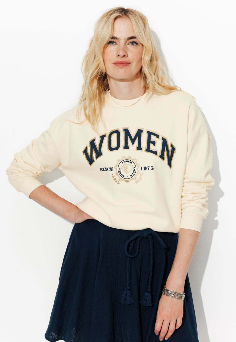 Femme blonde avec les cheveux lâches portant un sweat crème avec le texte "WOMEN", une jupe marine avec une ceinture tressée et un bracelet, posant devant un fond blanc.