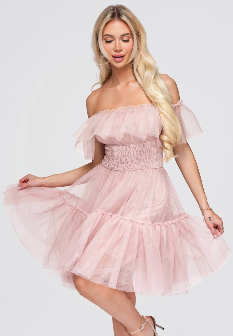 Femme aux longs cheveux blonds portant une robe en tulle rose clair à épaules dénudées, tenant la jupe déployée, debout devant un fond clair uni.