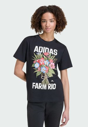 Frau mit lockigem Haar, die ein schwarzes Adidas-T-Shirt mit farbenfrohem Blumenstrauß-Grafik und schwarze Hose vor hellem Hintergrund trägt.