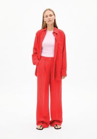 Rode linnen shirt met lange mouwen en knoopsluiting, gecombineerd met hoge rode linnen broek. Daaronder een nauwsluitend roze top. Zwarte flip-flops.