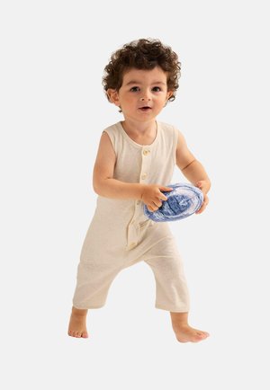 Hellbeige ärmelloser Jumpsuit mit Knöpfen und ausgestellten Beinen. Das Kind hält ein blaues Stofftier und steht barfuß vor einem weißen Hintergrund.