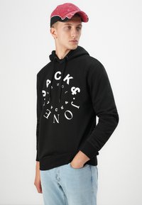 Musthave musta kapuutsiga, millel on ringikujuline valge graafiline tekst, varustatud nööridega kapuutsi ja soonikuga mansettidega, kombinatsioonis helesiniste teksade ja roosa mütsiga.