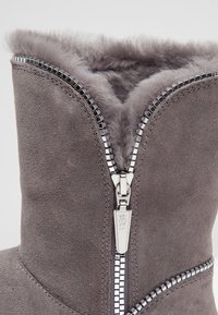 Šedá semišová bota se zipem s stříbrnými doplňky a plyšovou podšívkou. Design zahrnuje ozdobné šití kolem zipu.