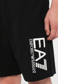 Short noir avec logo EA7 Emporio Armani blanc brodé sur la cuisse gauche, montré avec la main droite d'une personne posée à proximité.