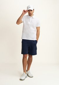Polo shirt bianco a maniche corte, pantaloni cargo blu navy e sneakers grigie chiare. Un cappellino bianco con logo nero. Look atletico semplice e casual.