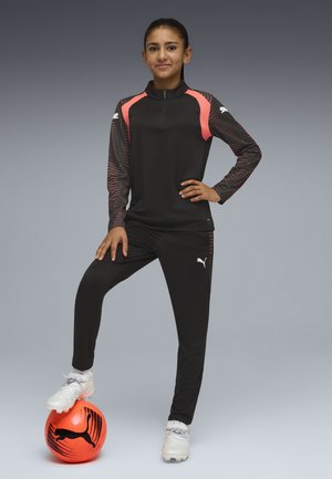 Joven jugadora de fútbol con chándal negro y coral descansando el pie sobre un balón de fútbol Puma naranja brillante contra un fondo gris.
