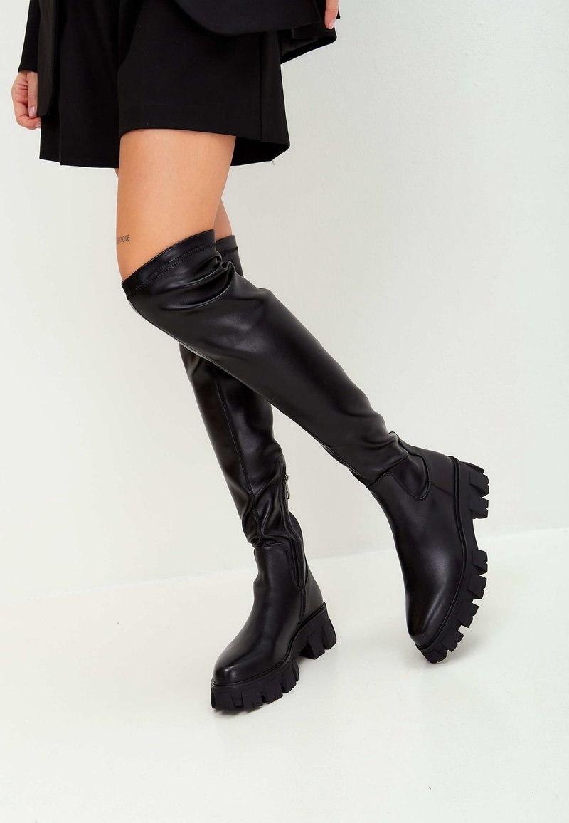 Botas de cuero negras hasta la rodilla con suela gruesa, que cuentan con una cremallera en el lado y una parte superior arrugada. Textura suave y diseño moderno.