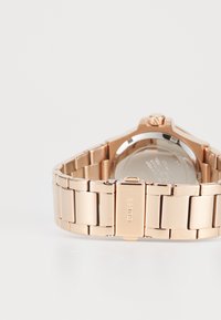 Montre en métal rose gold avec un bracelet à maillons, présentant un boîtier rond et un dos poli. Fermeture marquée "GUESS." Design minimaliste.